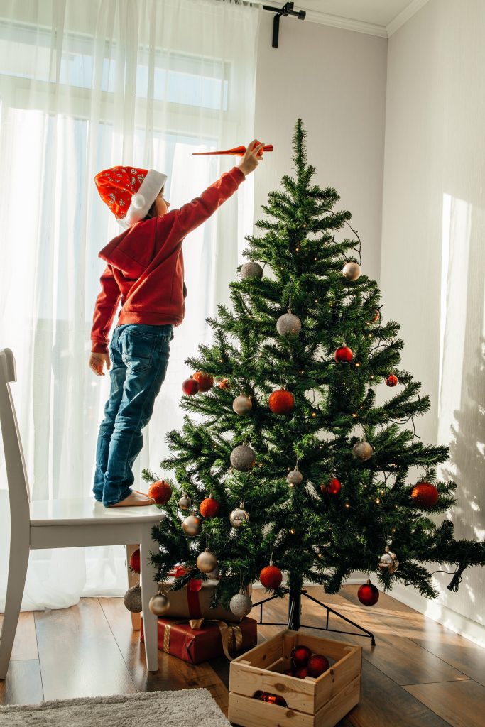 A boy in a santa claus hat decorates the New Year tree at home, puts a Christmas toy on the top of the tree. Children are preparing to celebrate Christmas and New Year